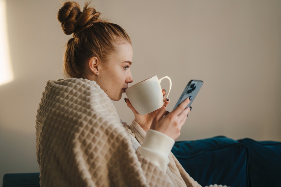 kobieta owinięta w koc trzyma telefon i kubek z herbatą