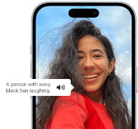 iPhone 15 z pokazaną funkcją VoiceOver, za pomocą której odczytywana jest informacja o obrazie; widać śmiejącą się osobę o czarnych, falowanych włosach